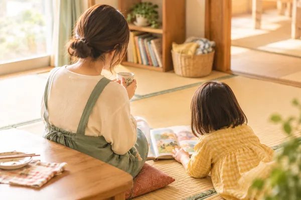 畳の部屋で、お茶を飲むお母さんと絵本を読む子どもの後ろ姿