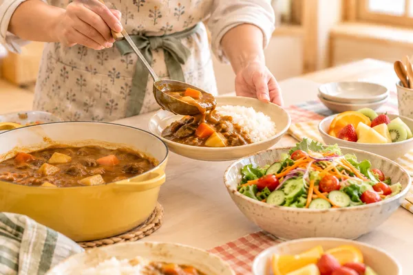 エプロン姿の大人が、カレーライスをお皿によそっている手元。横にサラダと果物